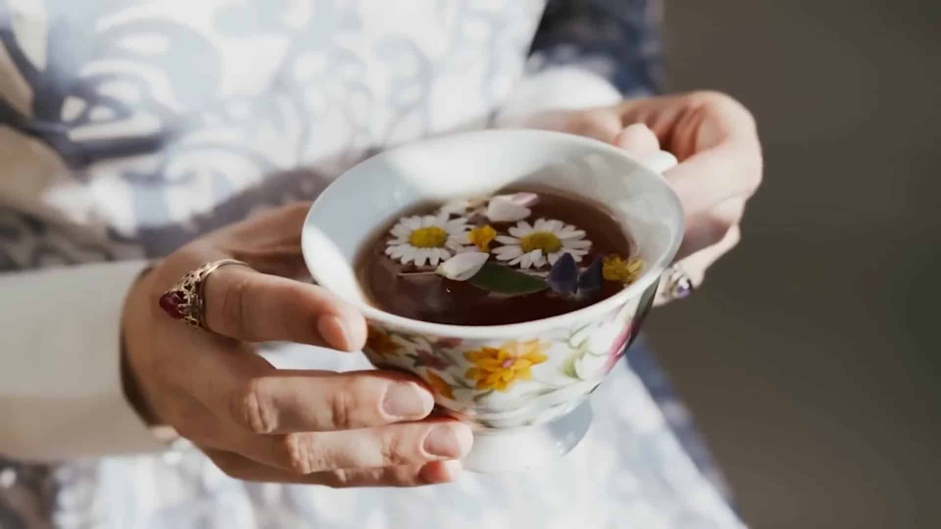 Person holding a floral teacup filled with tea, topped with floating chamomile flowers and petals, with a decorative ring visible on one finger.